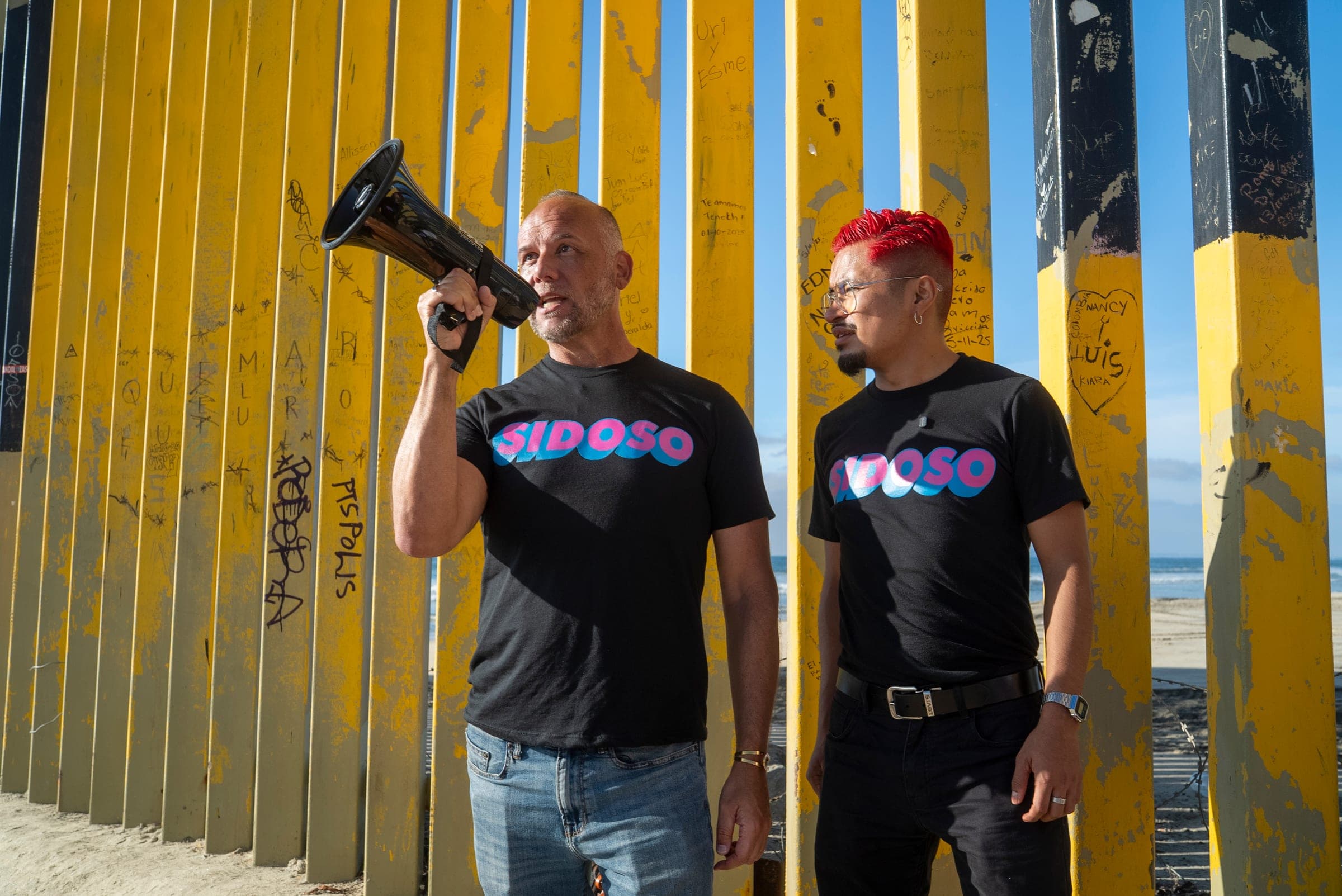 Two community advocates at a border wall, one speaking through a megaphone